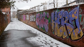 Alley snow This urban photograph captures Union Walk in Chesterfield, Derbyshire, United Kingdom, during a winter morning. The main subject is the alleyway, which is partially covered with snow, displaying a series of footprints along its length. The scene features brick architecture with graffiti art prominently visible on the walls, creating a vibrant contrast against the snowy environment. Snow has settled on the ground and on top of the walls, indicating recent cold weather. Barbed wire runs along the tops of the brick walls, typical of urban architectural security features. Residential buildings are visible in the background, highlighting the typical architecture found in this region of the United Kingdom. The image exemplifies the interplay between urban architecture and snow in a quiet Chesterfield alley.
